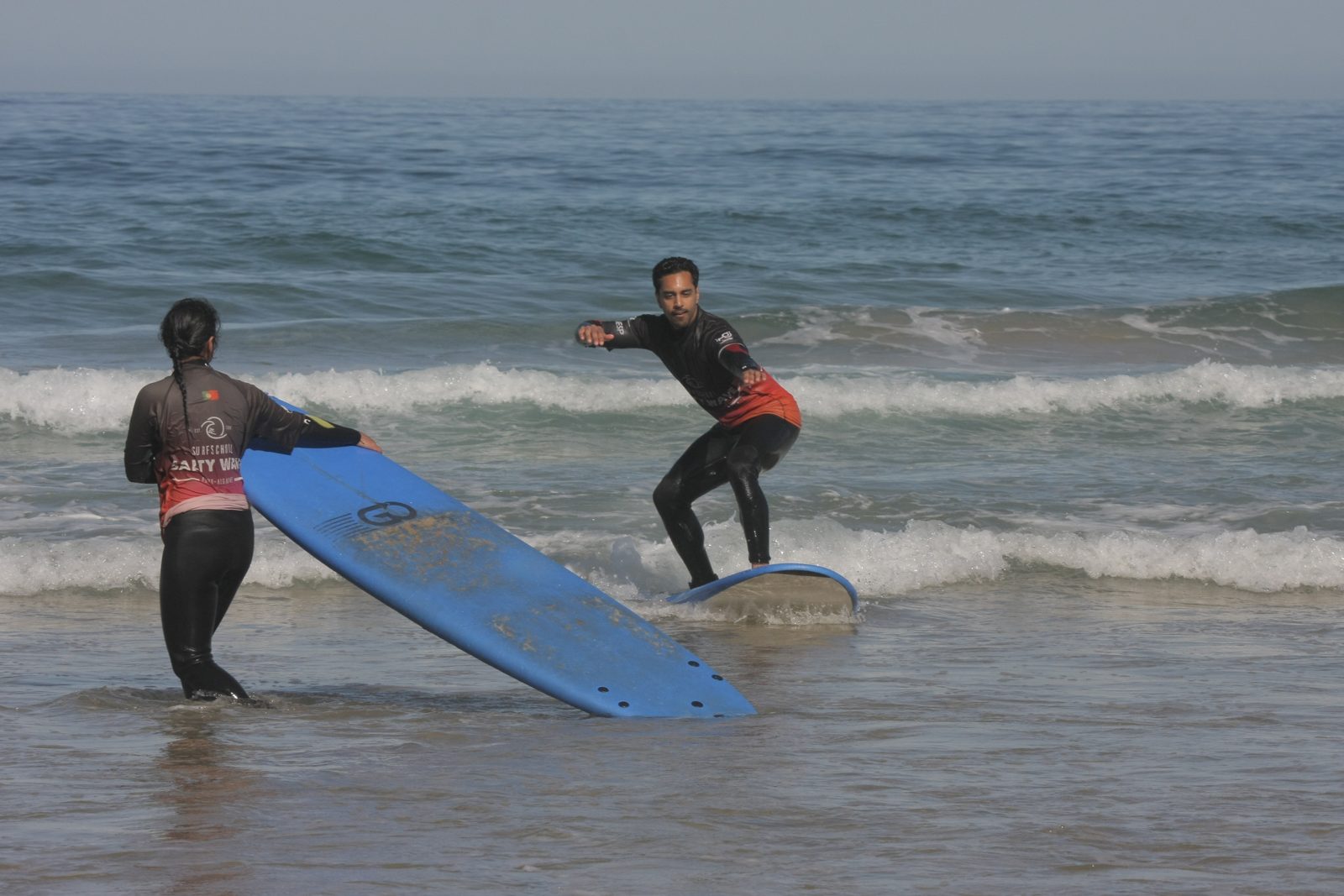 Beginner surfer standing up on his board during a surf lesson, with an instructor watching from the water